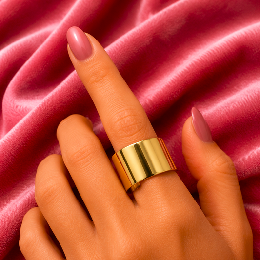 A close-up of a hand wearing a wide, polished gold ring, resting against a draped dusty pink velvet fabric background.