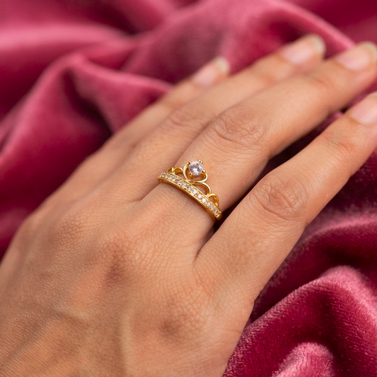 A gold ring with a heart-shaped clear gemstone and a pavé band, worn on a hand resting on a draped dusty rose velvet fabric.