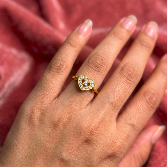 A gold ring with a shield-shaped pavé setting holding a small round center stone, worn on a hand against a pink velvet background.