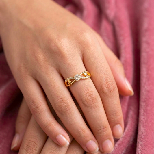 A close-up of a person's hands resting on a dusty rose velvet surface, wearing a delicate gold ring with a small cluster of clear stones on the ring finger.