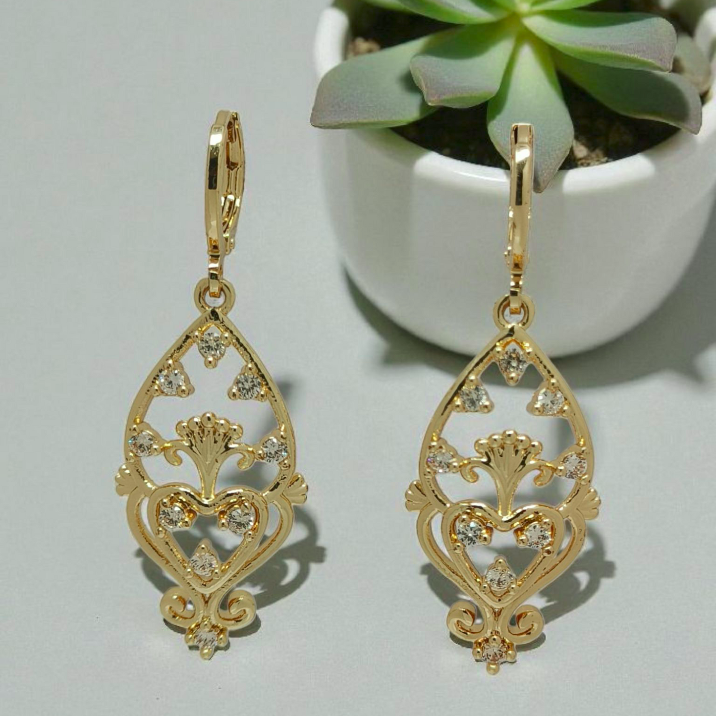 Gold earrings with intricate designs on a light gray background with a potted plant.