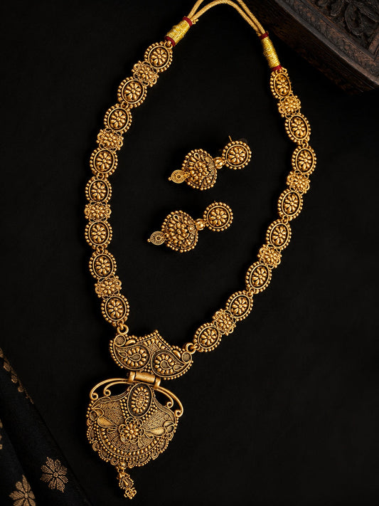 An ornate gold jewelry set featuring a necklace with a large decorative pendant, matching drop earrings, and a yellow cord, displayed on a dark black background.