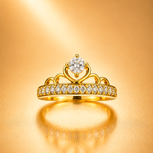 A gold crown-shaped ring with a round clear center stone and a row of small clear stones along the band, shown on a warm gold background with a soft reflection below.