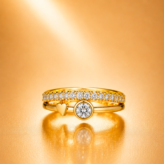 A gold ring set with a row of small clear stones and a central round clear stone, accented by a small gold heart, photographed on a warm gold reflective surface.