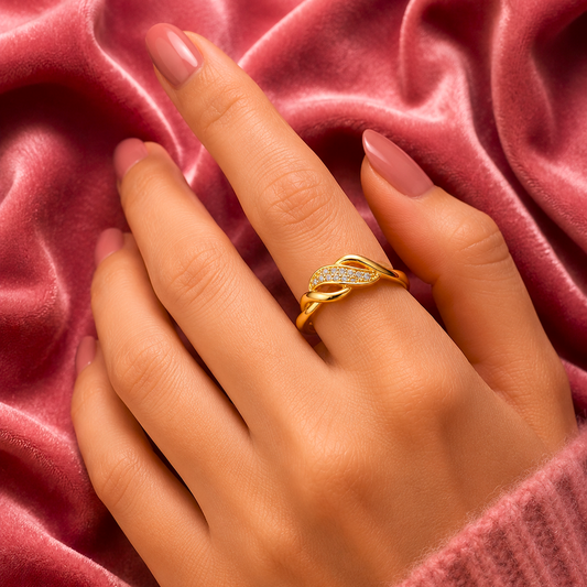 A gold ring with a twisted leaf design set with small clear stones, worn on a hand with pink manicured nails, resting on a draped dusty pink velvet fabric.