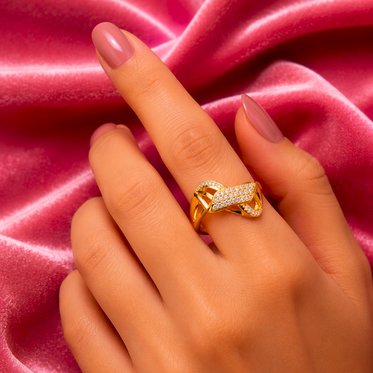 A close-up of a hand wearing a gold ring with pavé-set clear stones, posed against a draped pink satin fabric background.
