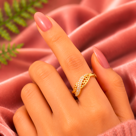 A gold ring with a textured openwork band and small clear stones, shown worn on a hand with pink manicured nails against a pink satin fabric background.