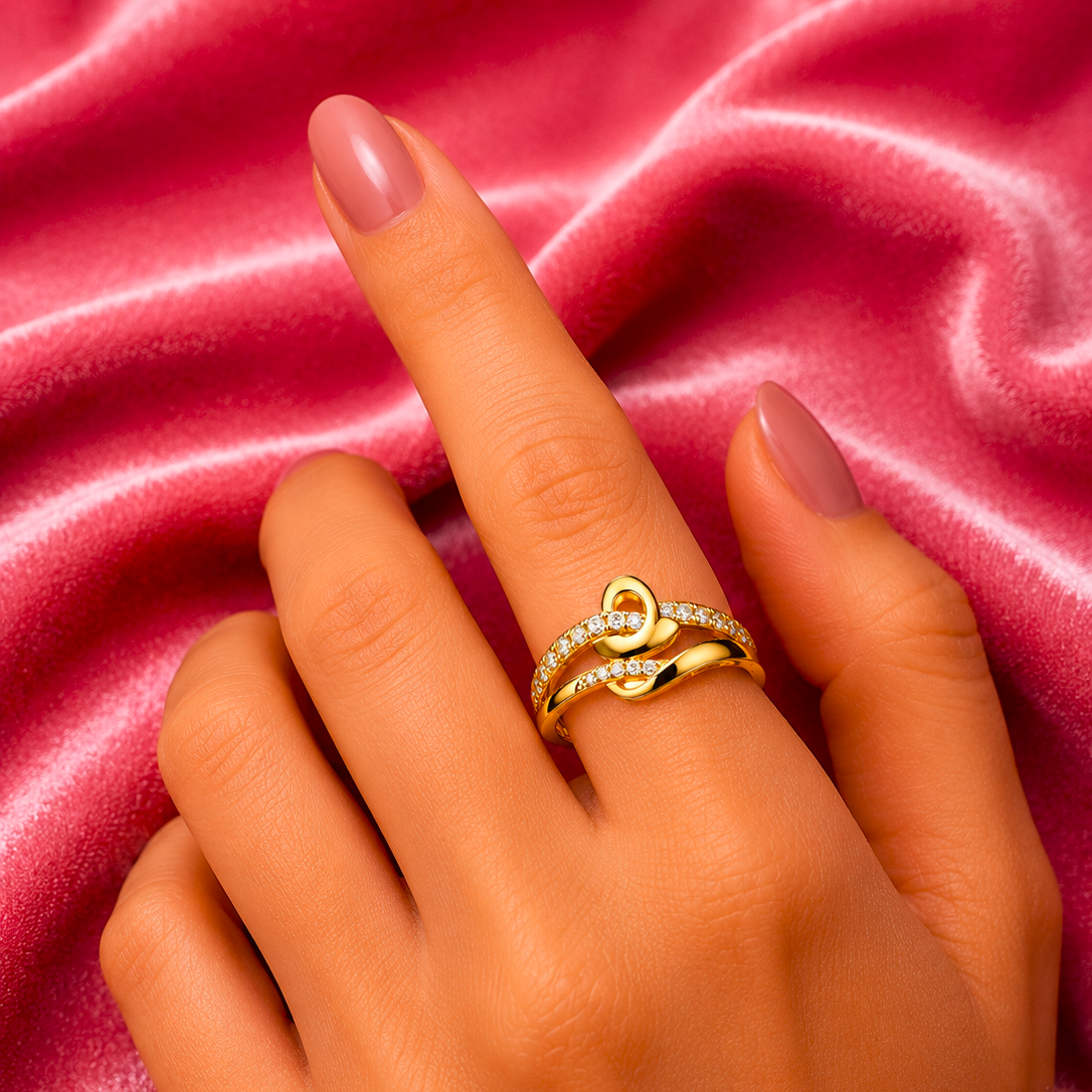 A close-up of a hand wearing a gold ring with a heart-shaped design and small clear stones, set against a pink satin fabric background.