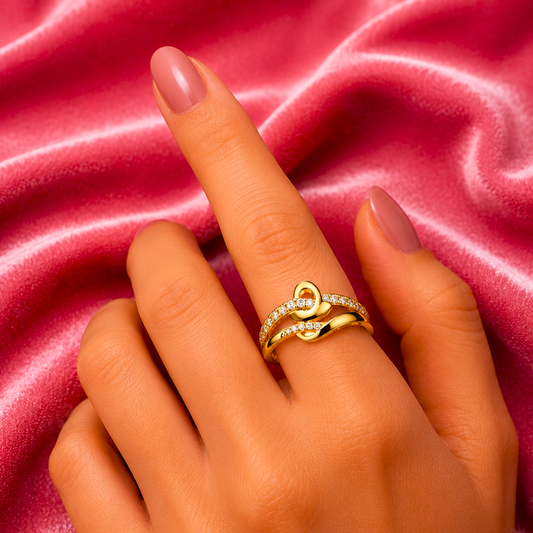 A close-up of a hand wearing a gold ring with a heart-shaped design and small clear stones, set against a pink satin fabric background.