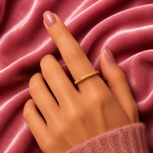 A close-up of a hand wearing a delicate gold ring with engraved text, resting against a draped pink velvet fabric background.