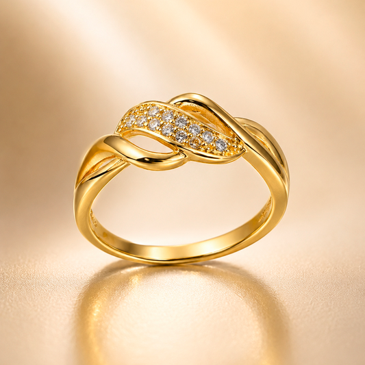 A gold ring with a twisted knot design and a pavé-set row of clear stones across the top, displayed on a warm beige satin background.
