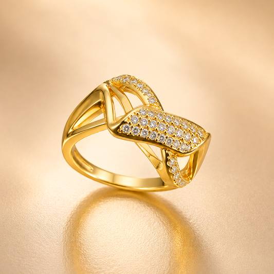 A gold ring with a geometric, ribbon-like design paved with small clear stones, displayed on a warm, reflective beige surface.