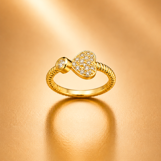 A gold ring featuring a heart-shaped pavé center with a small round clear stone on one side and a textured rope-style band, photographed on a warm gold reflective surface.