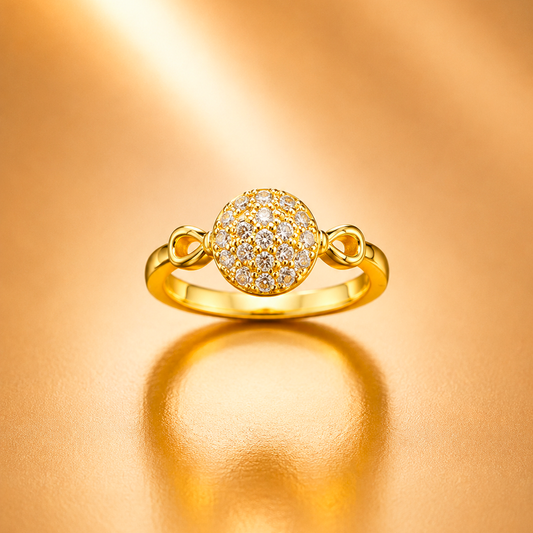 A gold ring featuring a round pavé-set cluster of small clear stones in the center, with decorative side loops on a polished gold band, displayed on a warm gold reflective surface.