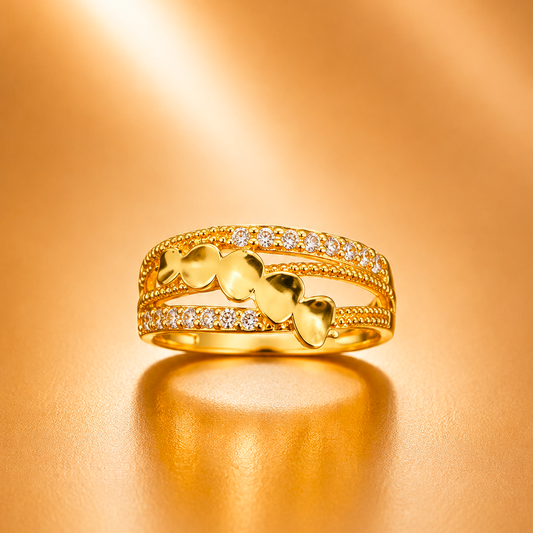 A gold ring with three bands, featuring three heart-shaped charms in the center, a row of small clear stones on the upper and lower bands, and a textured rope-like detail, displayed on a warm gold background.