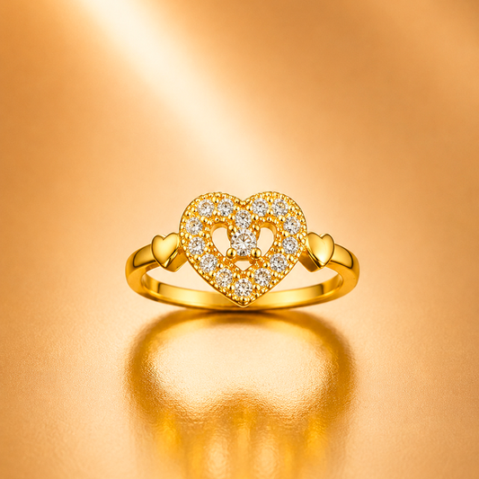 A gold heart-shaped ring with pavé-set clear stones and a central letter V, displayed on a warm, reflective gold surface.