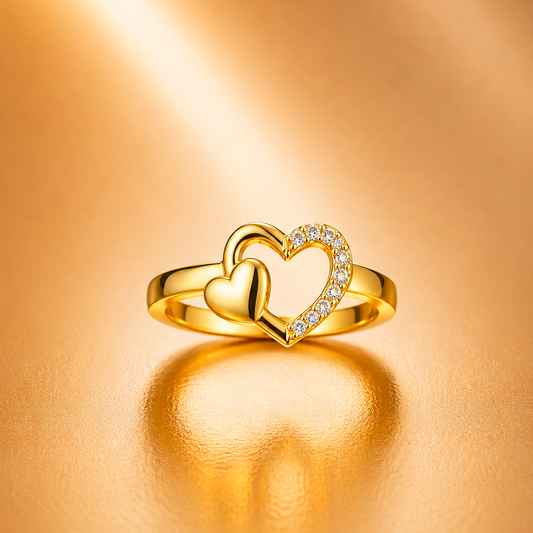 A gold ring featuring two interlocking heart shapes, with one heart adorned with small clear stones, set against a warm, reflective gold background.