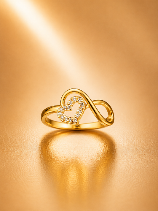A gold infinity loop ring featuring a central open heart paved with small clear stones, displayed on a warm, reflective gold surface.