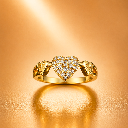 A gold ring featuring a central pavé heart flanked by two smaller hearts, photographed on a warm metallic gold background with a soft reflection below.