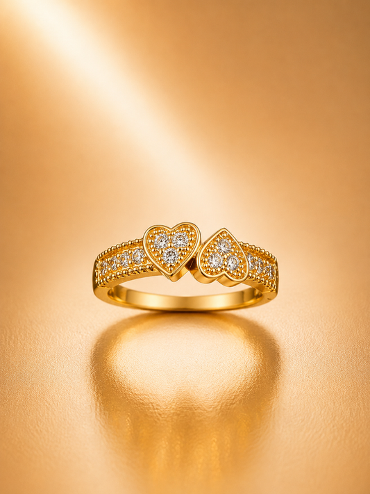 A gold ring featuring two heart-shaped pavé settings with small clear stones, shown on a warm gold reflective surface.
