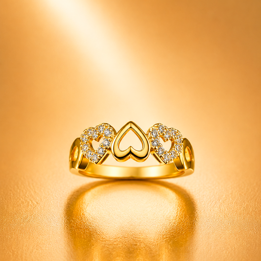 A gold ring with three heart-shaped motifs, including two pavé-set clear stones in the side hearts and a larger open heart in the center, displayed on a warm gold reflective surface.