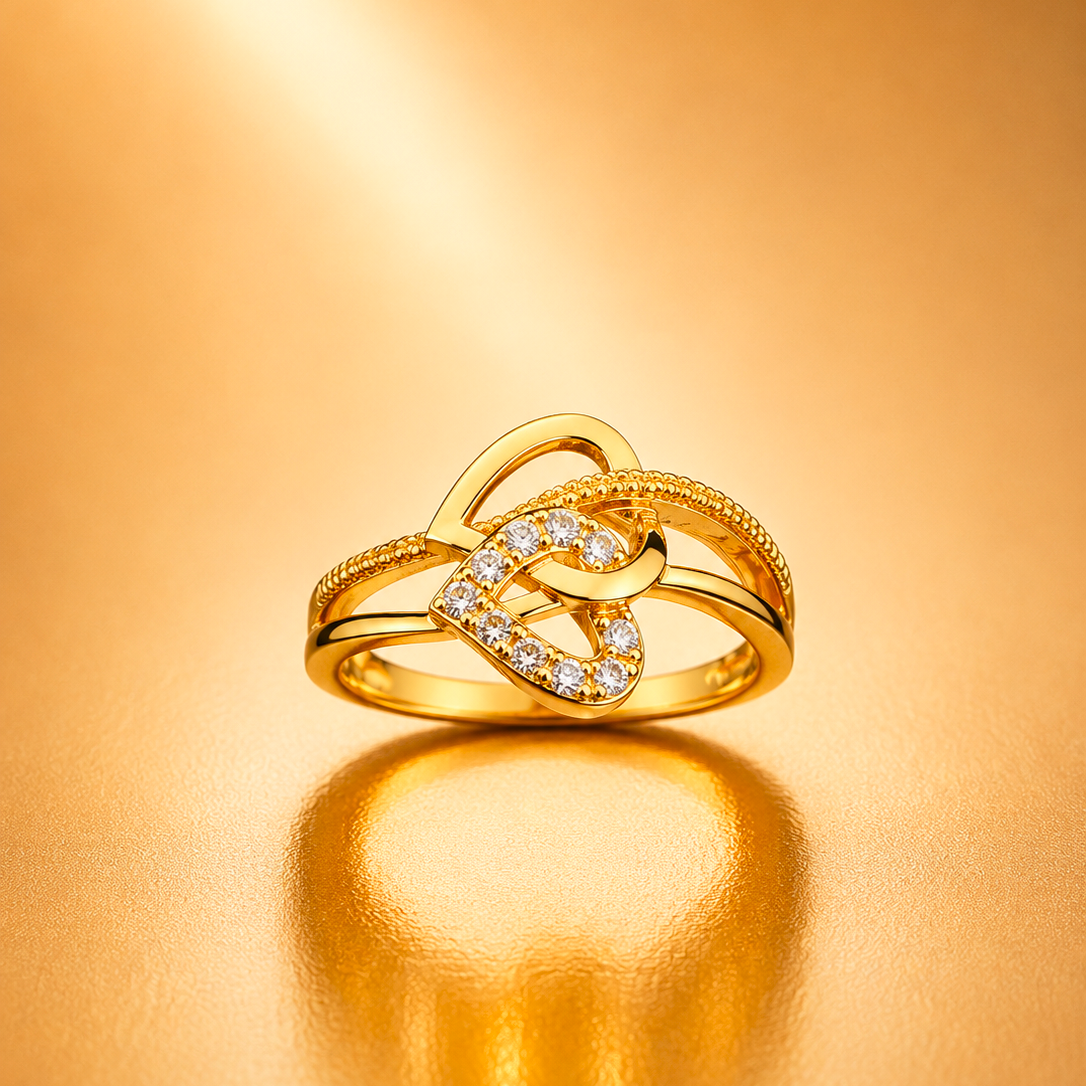 A gold ring with an intertwined knot design, featuring a pavé-set diamond heart in the center and textured beaded bands, displayed on a warm gold reflective surface.