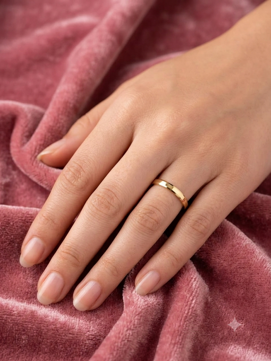 A close-up of a person's hand resting on a dusty rose velvet blanket, wearing a simple, polished gold band ring.