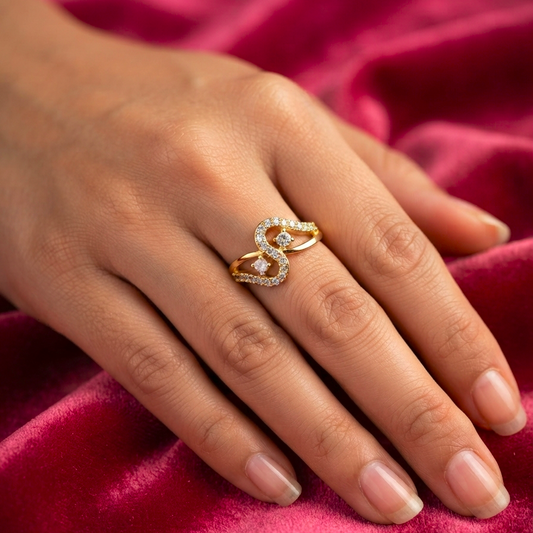 A close-up of a hand wearing a gold ring with a stylized interlocking double-S design set with small clear stones, resting on a draped deep pink velvet fabric.