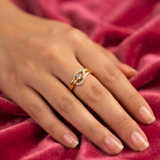 A gold ring with a central pink gemstone and small clear stones, worn on a hand resting on a textured pink velvet fabric.