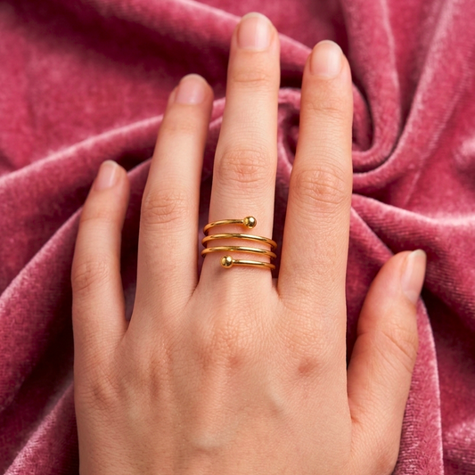 A close-up of a hand wearing three thin gold wrap rings with small bead details, set against a draped dusty rose velvet fabric.