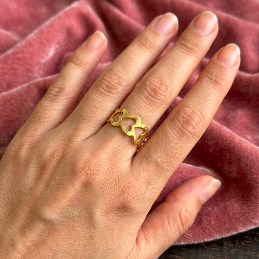 A gold ring with a heart-shaped cutout design worn on a person's ring finger, set against a soft, draped dusty rose velvet fabric background.