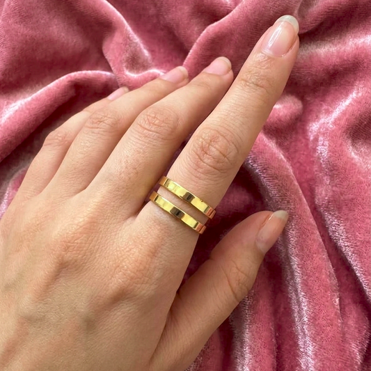 A close-up of a hand wearing two slim gold bands rings, resting on a textured, draped dusty pink velvet fabric.