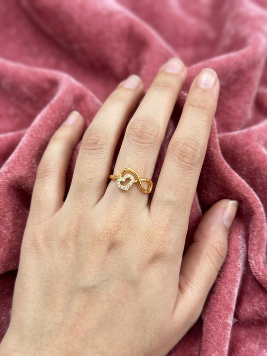 A gold infinity ring with small clear stones on the ring finger of a hand, resting on a textured dusty pink velvet fabric.