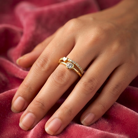 A close-up of a hand wearing a gold ring set with a round-cut center stone and a small heart-shaped stone, resting on a textured dusty rose velvet fabric.