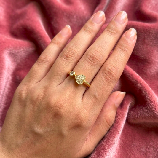 A gold ring with a heart-shaped pavé center and a small round accent stone, worn on a hand resting on a dusty rose velvet fabric.