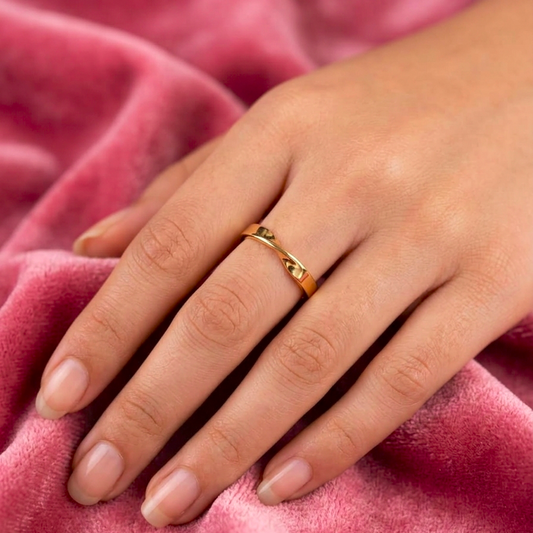 A close-up of a hand wearing a delicate gold ring with a small central stone, resting on a soft pink velvet fabric background.