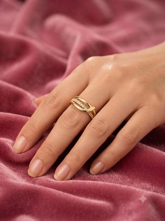 A close-up of a hand wearing a gold ring with a twisted, open design and small clear stones, resting on a textured pink velvet fabric.