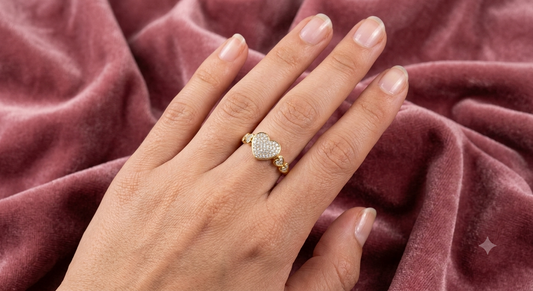 A gold ring with a heart-shaped pavé center and small side accents, worn on a hand resting on a textured dusty rose velvet fabric.