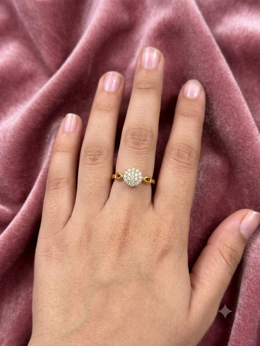 A gold ring with a round cluster of clear pavé stones worn on a person's ring finger, set against a draped dusty rose velvet fabric background.