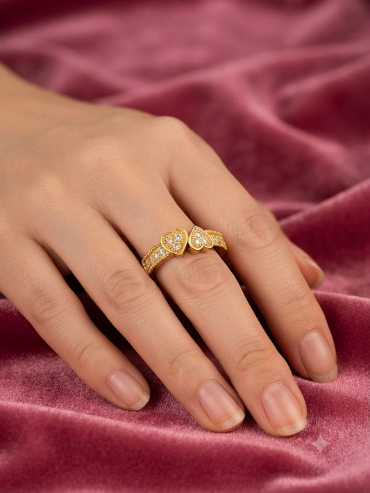 A close-up of a hand wearing a gold ring with two heart-shaped pavé settings, resting on a draped dusty rose velvet fabric.