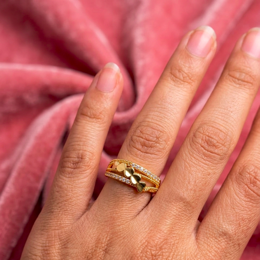 A gold ring with a small heart-shaped charm and a row of clear stones, worn on a person's finger against a pink velvet background.