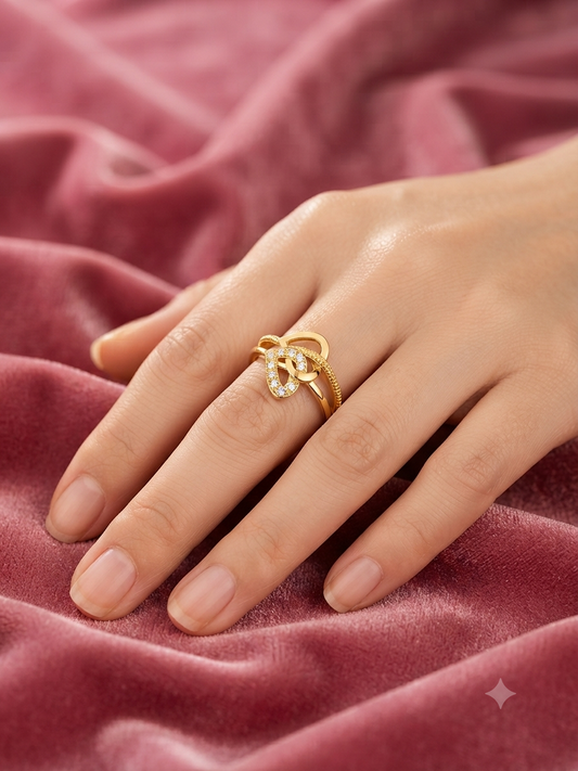 A gold ring with a bow-like knot design and small clear stones, worn on a hand resting on a dusty pink velvet fabric background.