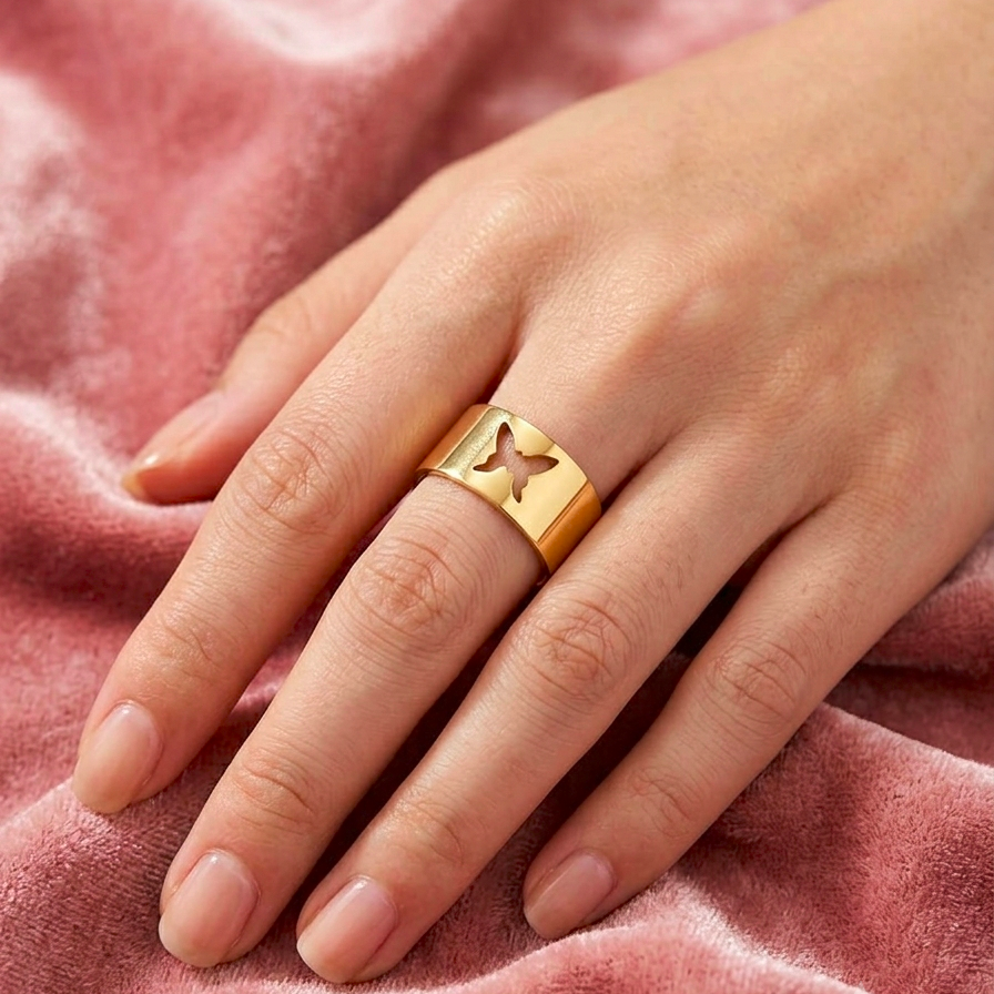 A gold ring with a cutout butterfly design on the side, worn on a person's ring finger over a soft pink velvet fabric background.