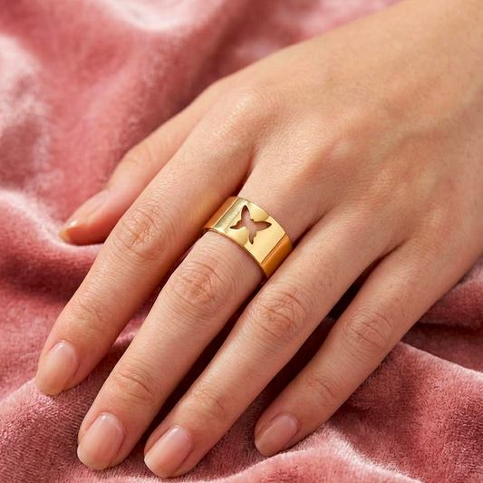 A gold ring with a cutout butterfly design on the side, worn on a person's ring finger over a soft pink velvet fabric background.