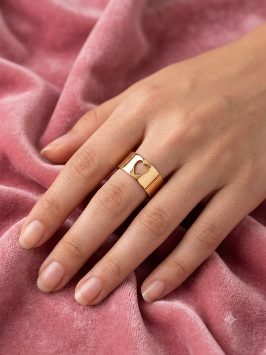 A gold ring with a heart-shaped cutout, worn on a hand resting on a soft pink velvet fabric.