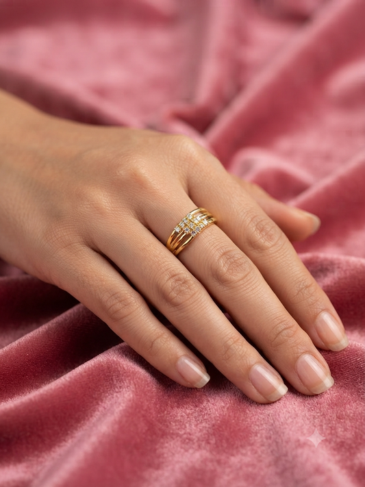 A close-up of a hand resting on a pink velvet surface, wearing a delicate gold ring with small clear stones.