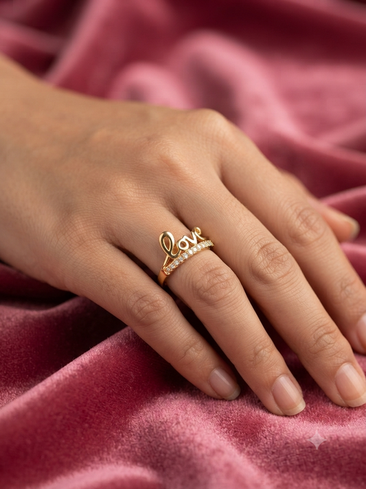 A gold ring with the word 'Love' in cursive and small clear stones, worn on a person's finger against a pink velvet background.