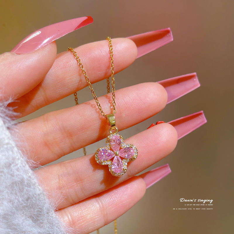 Hand holding a gold necklace with a pink flower pendant against a blurred background