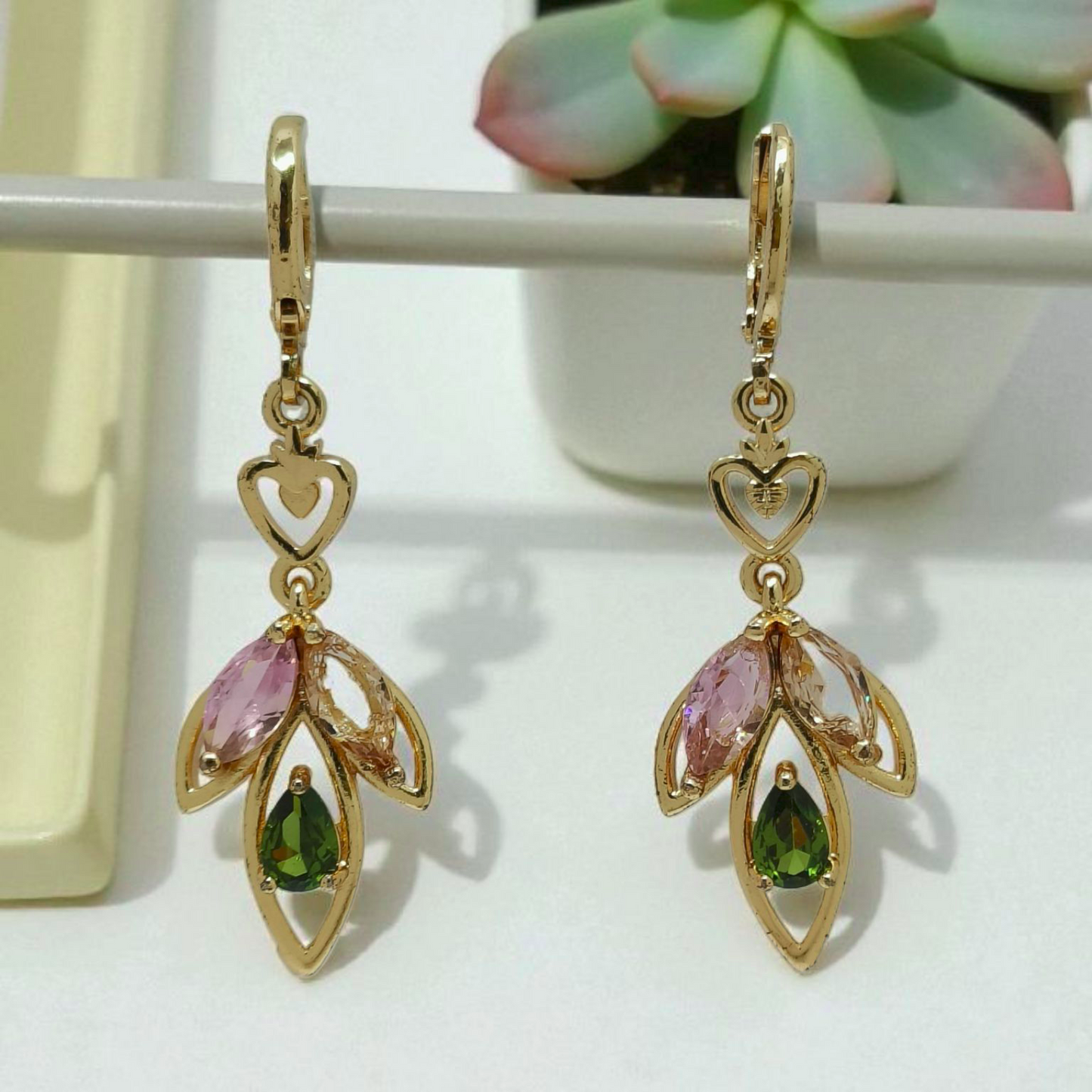 Gold earrings with pink and green gemstones on a white surface with a succulent plant in the background.