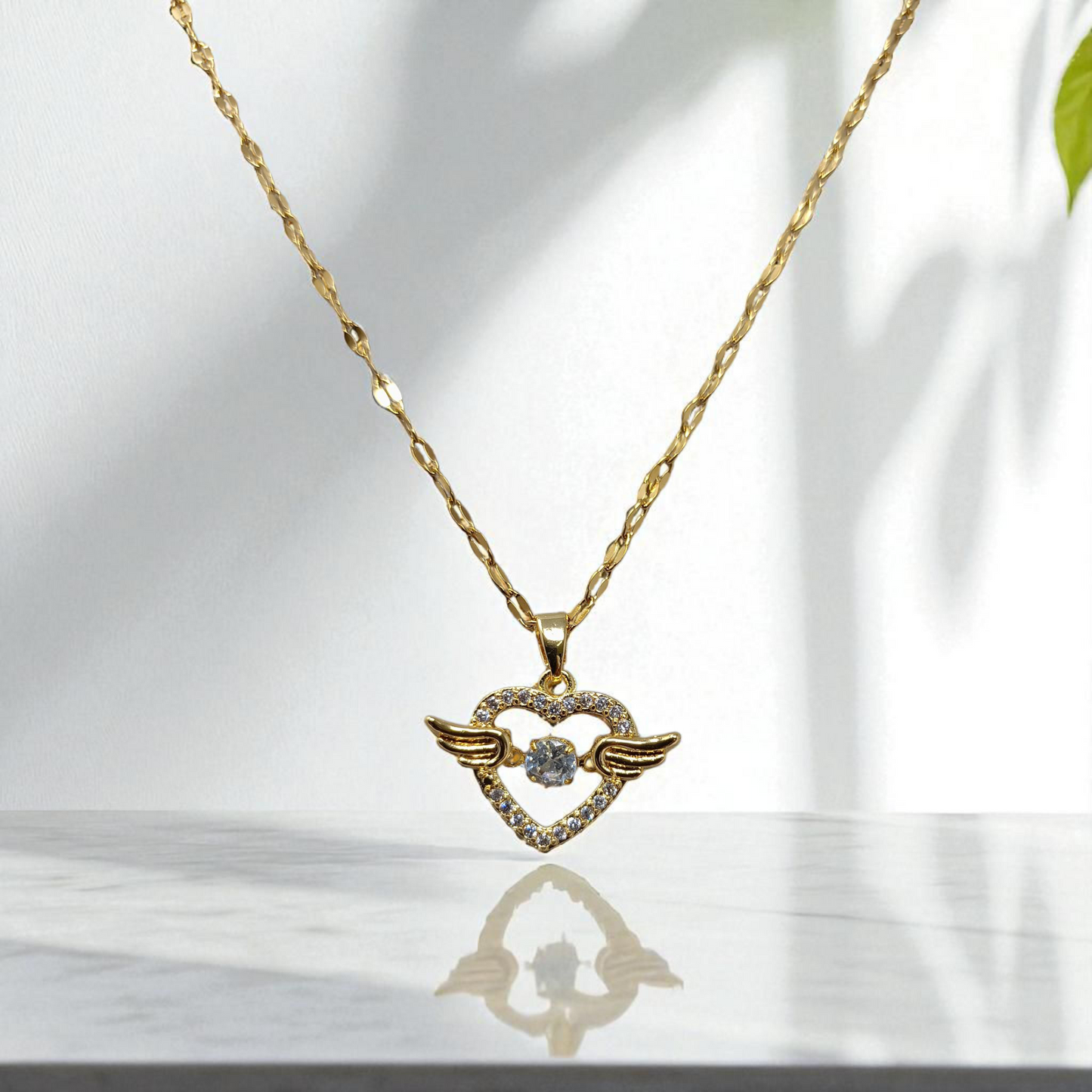 Gold necklace with a heart-shaped pendant featuring wings on a reflective surface.
